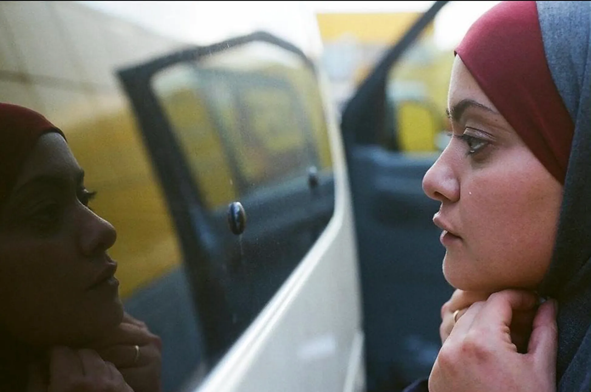 A woman in a hijab adjusts her scarf while studying her reflection in a car window
