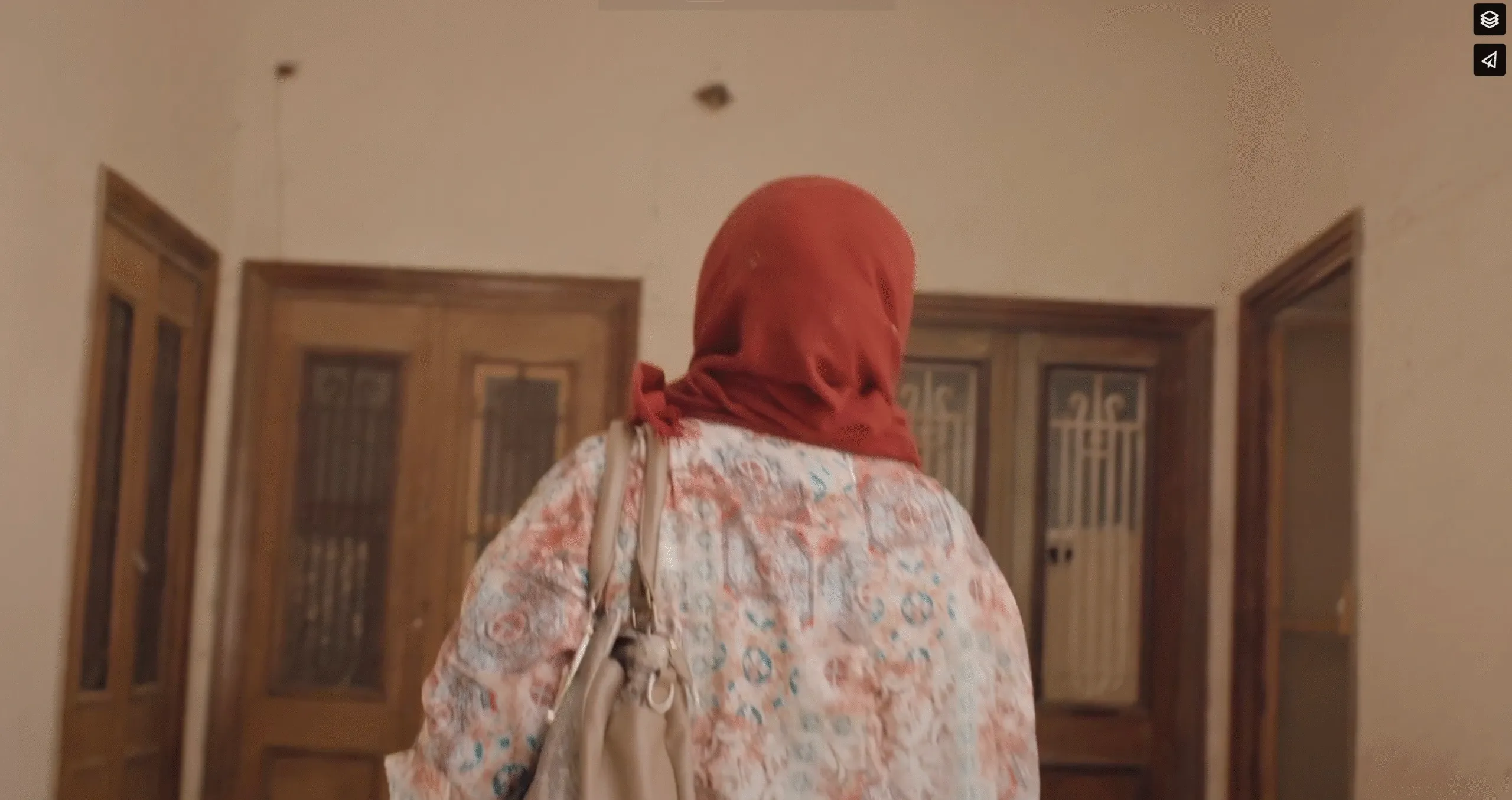 A woman in a red hijab and pastel blouse walks through a sunlit corridor of wooden doors